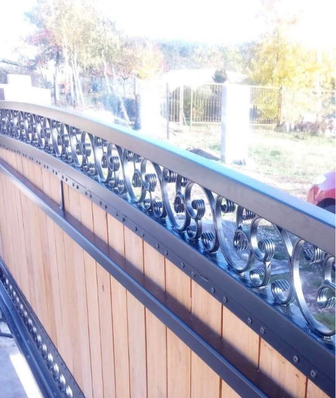 Drewniano-metalowa brama wjazdowa z dekoracyjnymi, kutymi elementami i nitowanymi detalami, widziana pod kątem, z fragmentem ogrodzenia w tle.