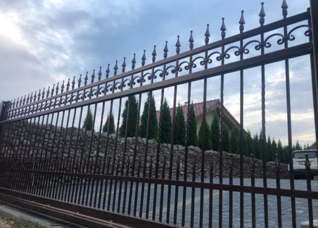 Ciemnobrązowa brama wjazdowa z dekoracyjnymi grotami, na tle kamiennego muru, zielonych drzew i fragmentu domu z czerwoną dachówką. Pochmurne niebo.