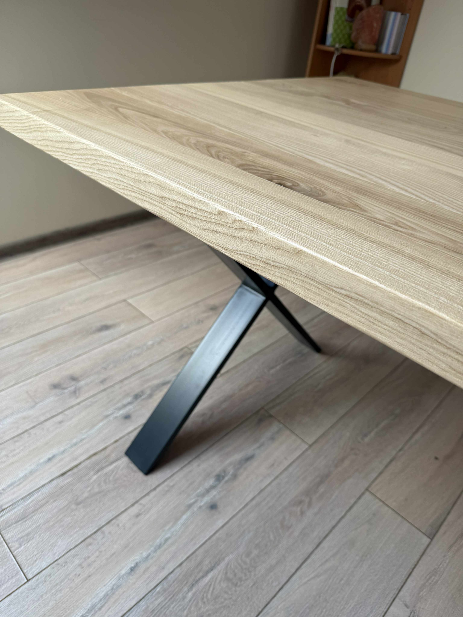 Stół z blatem z jasnego drewna i czarną, metalową nogą w kształcie litery X, ustawiony na podłodze z paneli. Widok z niskiego kąta, ukazujący strukturę drewna.