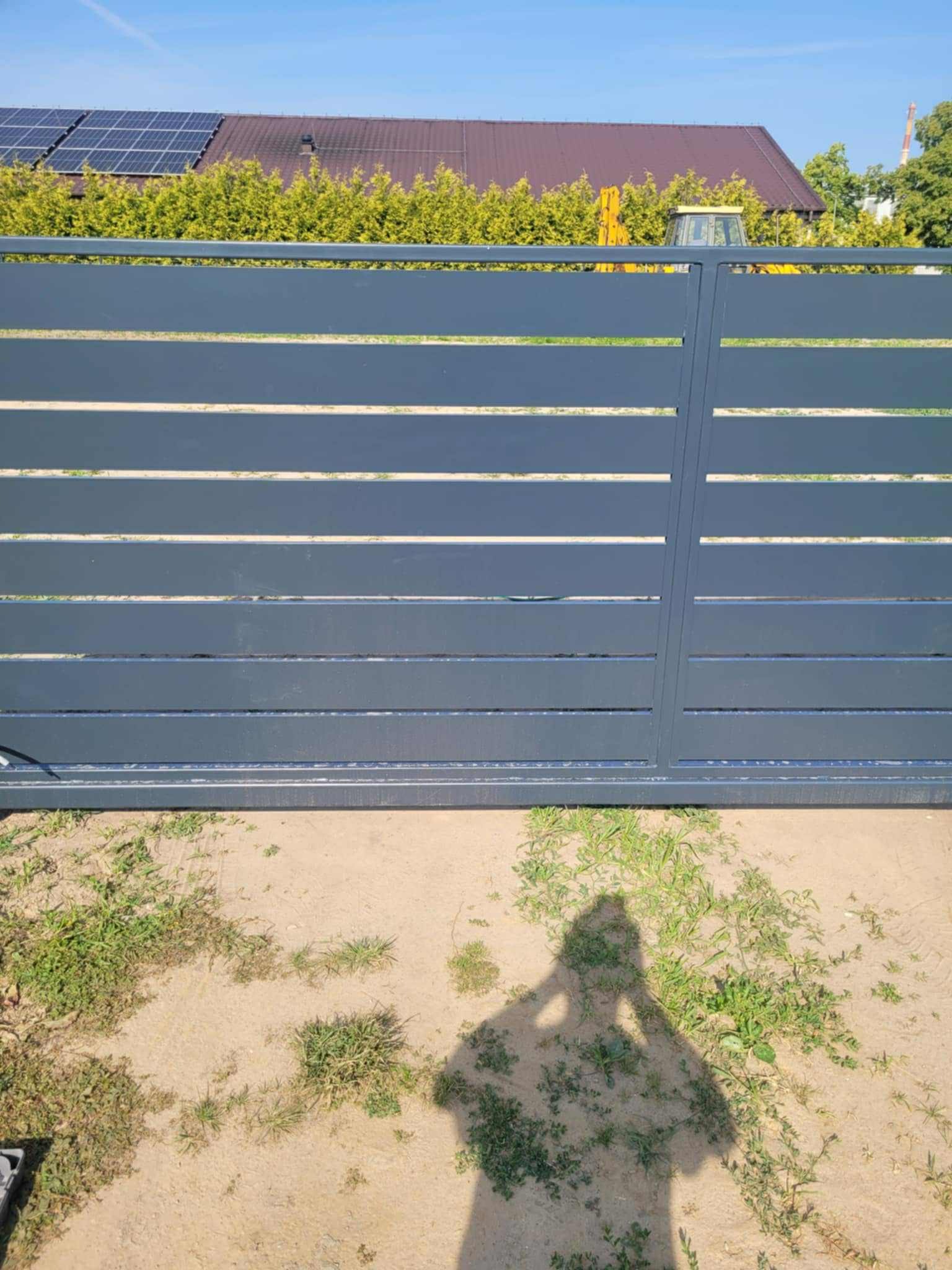 Nowoczesna, pozioma brama wjazdowa w kolorze grafitowym, z widocznym cieniem osoby fotografującej na piaszczystym podłożu i zielenią w tle.