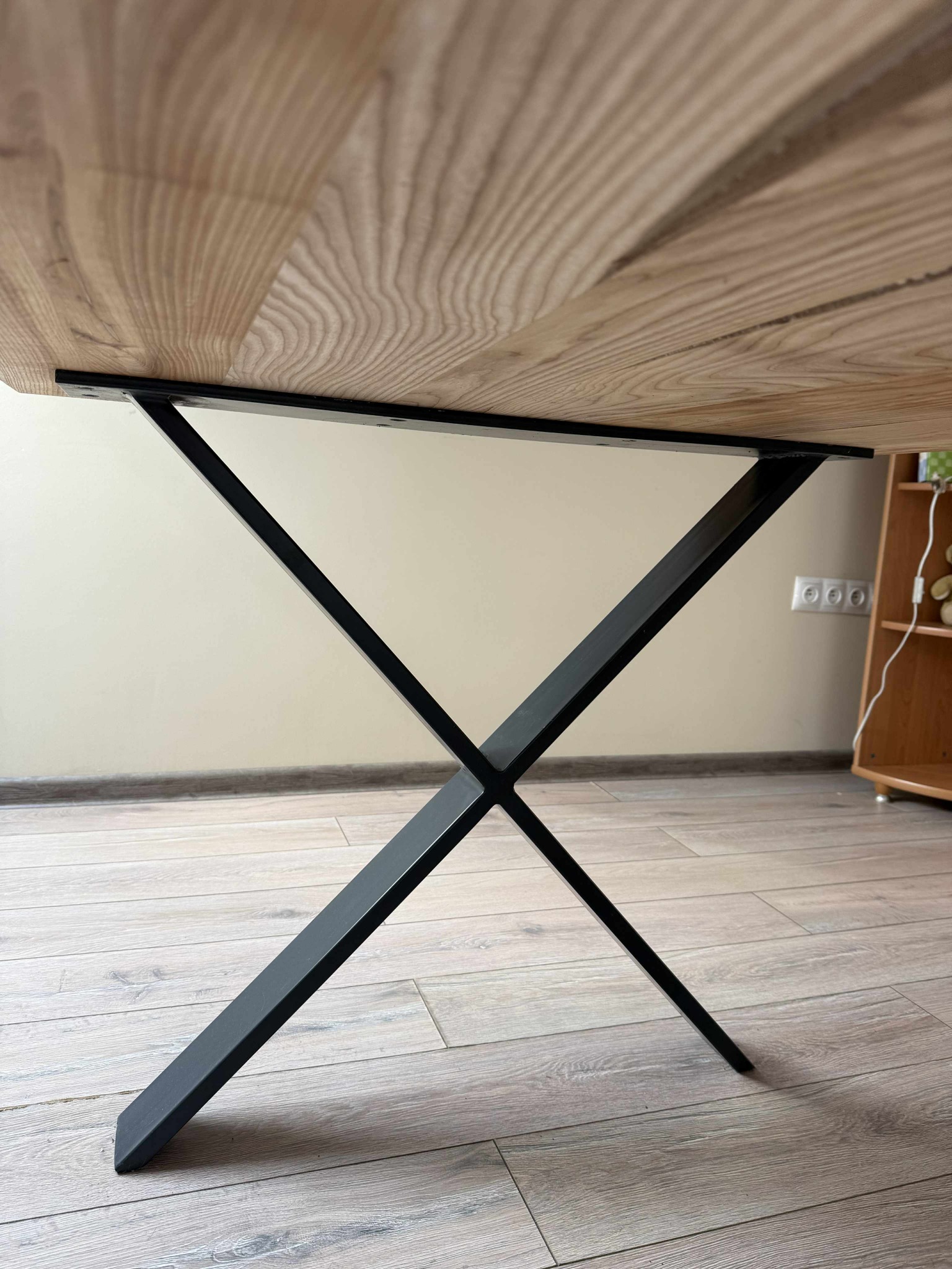 Stół z drewnianym blatem i metalową, czarną podstawą w kształcie litery X. Widok z dołu, ukazujący strukturę drewna i detale łączenia elementów.