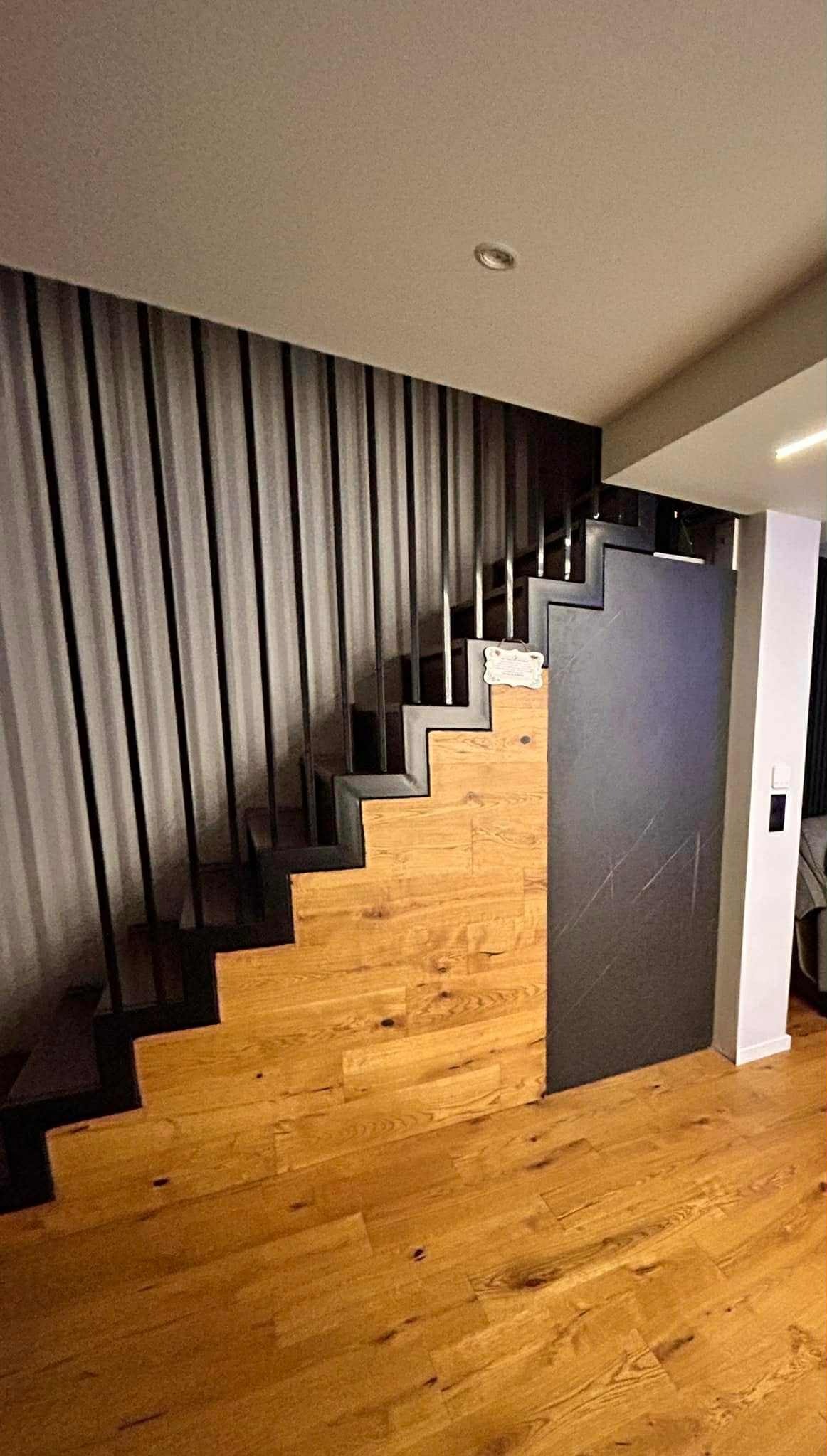 Nowoczesne schody z drewnianymi stopniami i czarną, metalową balustradą z pionowych prętów. Widoczna drewniana podłoga i fragment szarej ściany.