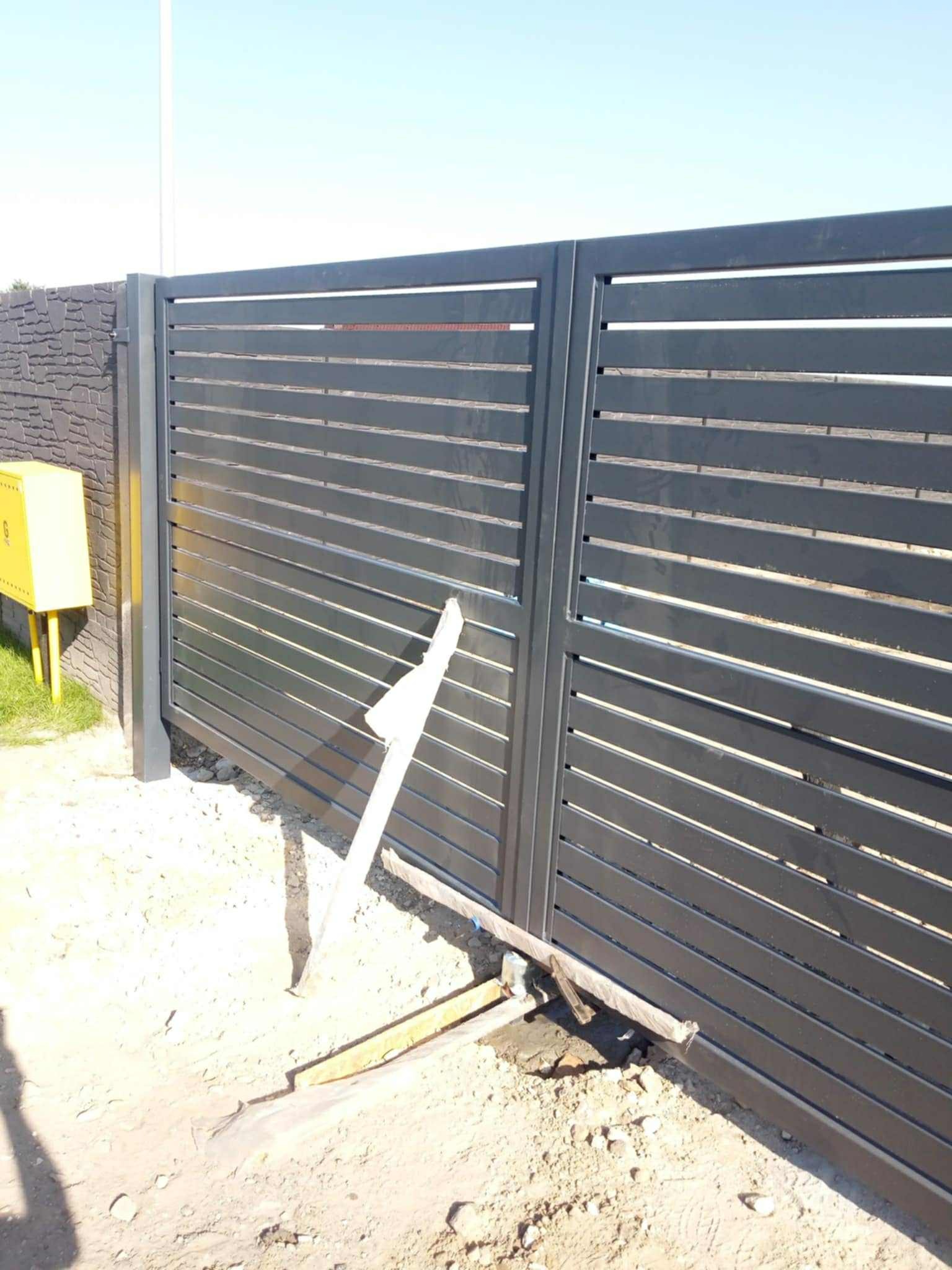 Nowoczesna, przesuwna brama wjazdowa z poziomymi panelami w kolorze grafitowym. Widoczne ślady montażu i żółta skrzynka gazowa obok ogrodzenia.