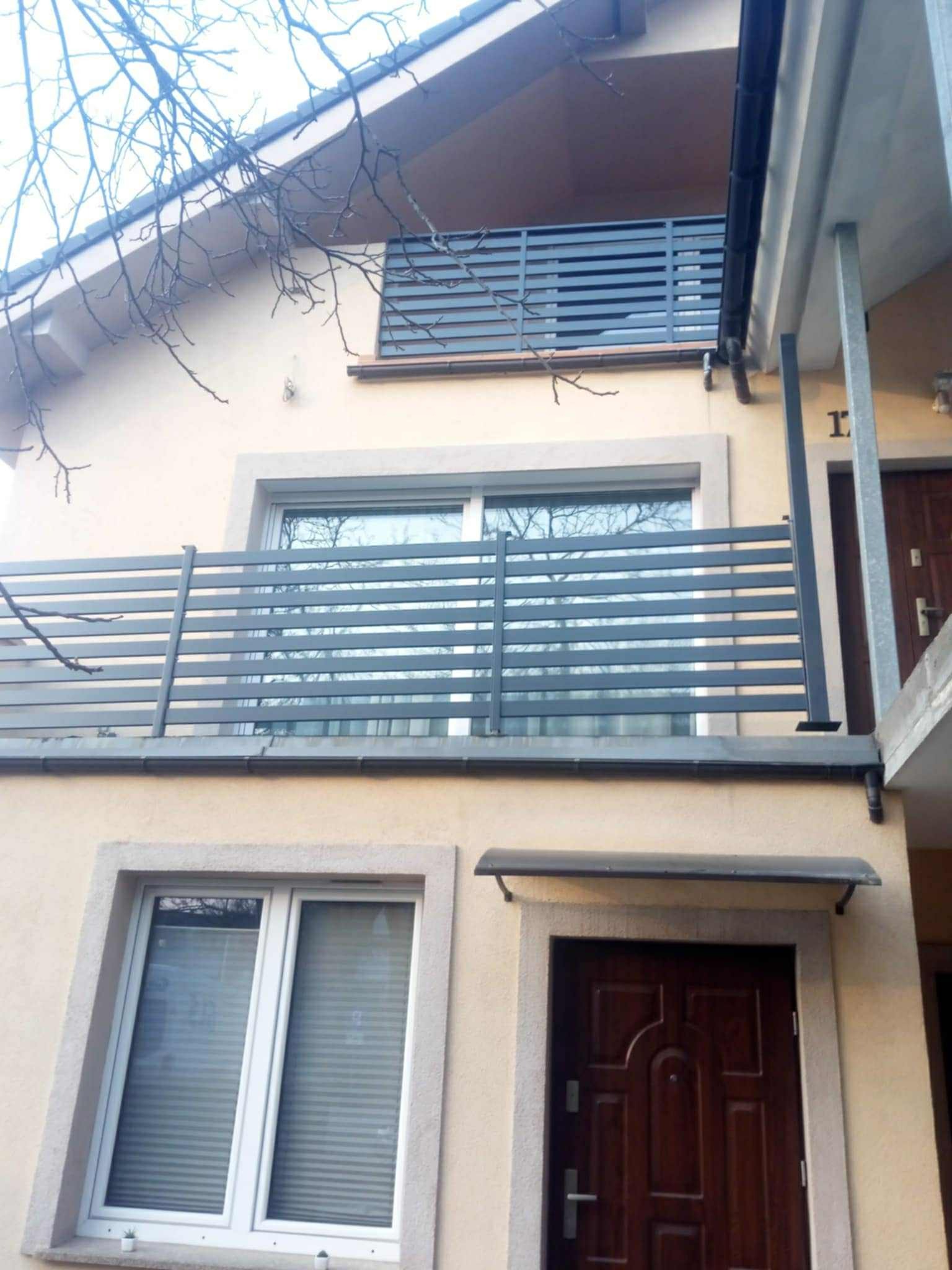 Nowoczesna, pozioma balustrada balkonowa z metalowych profili, szara, zamontowana na budynku o jasnej elewacji. Widok z dołu, fragment drzewa w kadrze.
