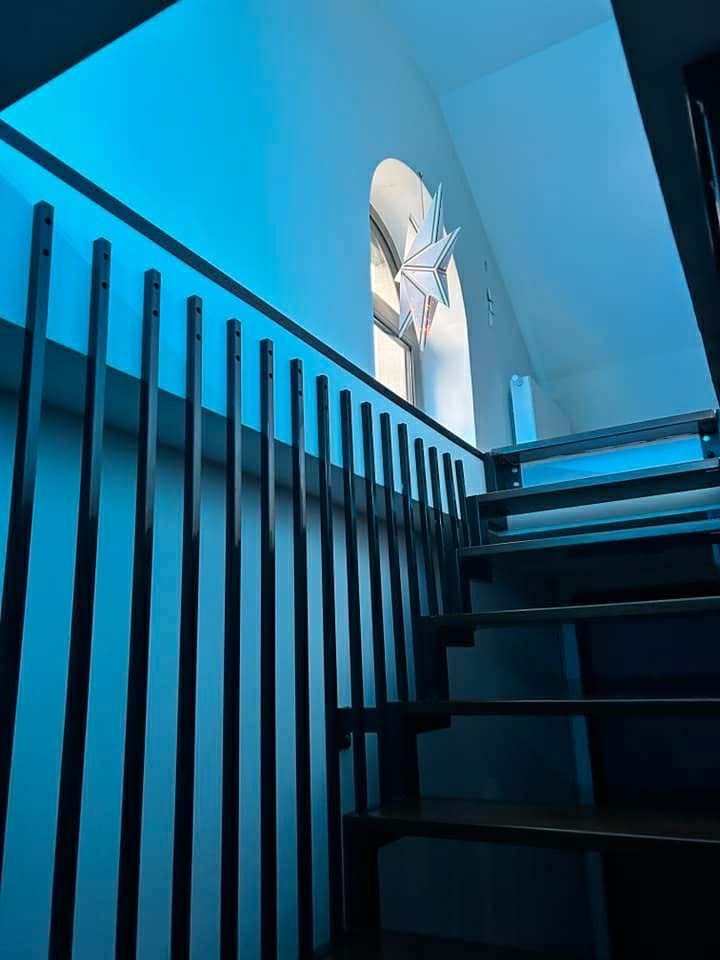 Nowoczesna balustrada schodowa z pionowych, czarnych prętów, oświetlona niebieskim światłem, z widocznym oknem łukowym i dekoracją w kształcie gwiazdy.