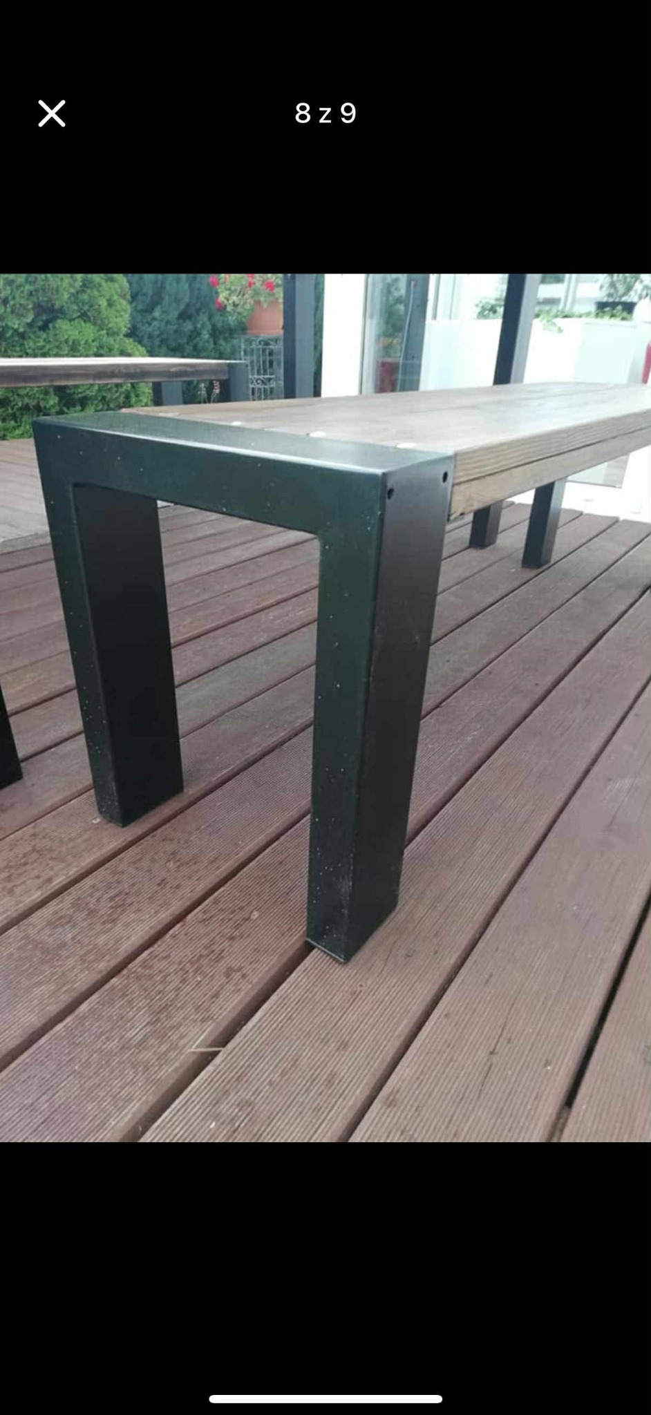 Metalowa noga stołu na drewnianym tarasie. Noga w kształcie litery U, pomalowana na czarno, mocowana do drewnianego blatu. Widoczne deski tarasowe i zieleń w tle.