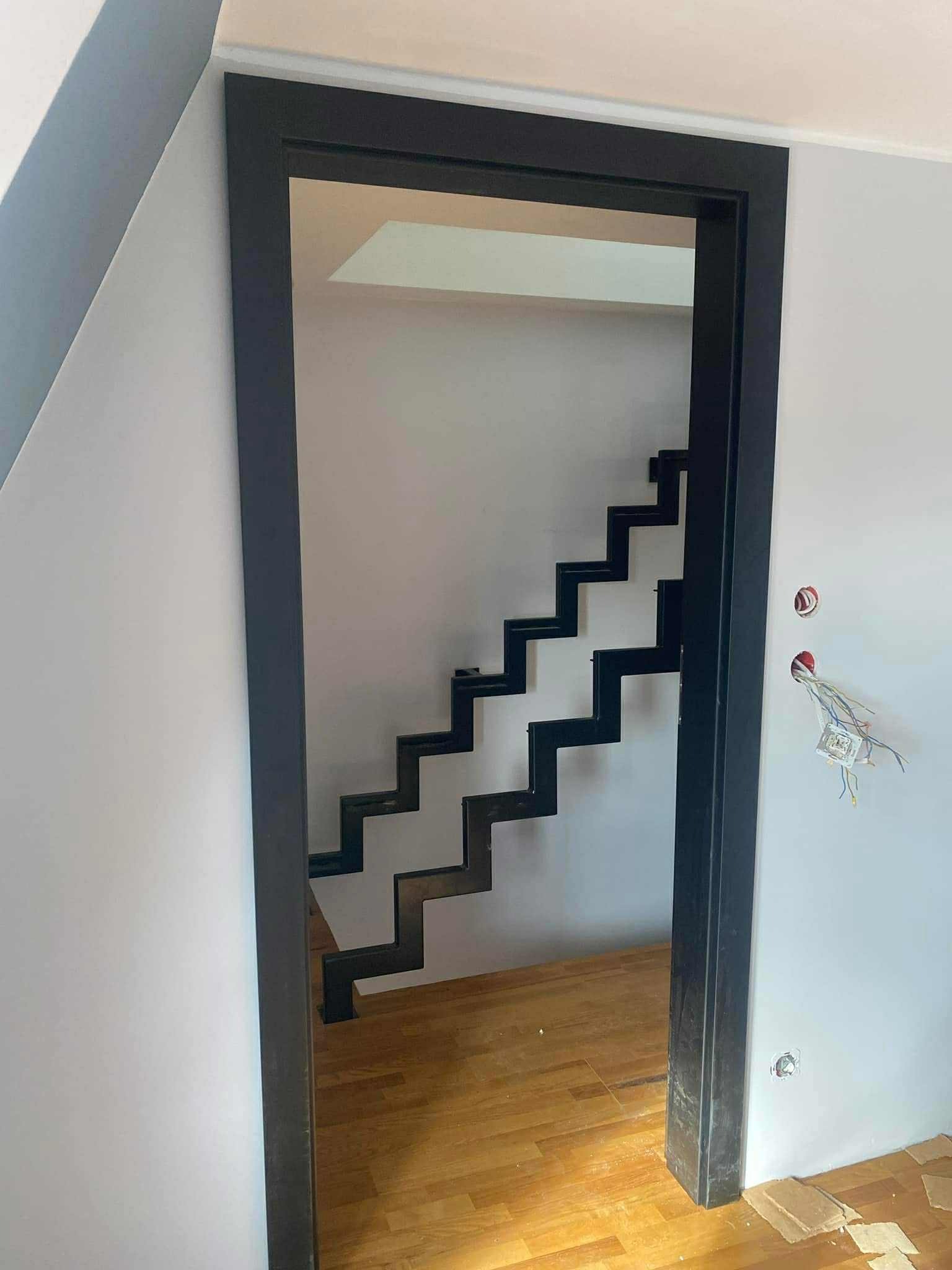 Nowoczesne, minimalistyczne schody metalowe w czarnej ramie, widoczne z perspektywy wejścia. Drewniana podłoga. Widoczne niedokończone prace elektryczne.