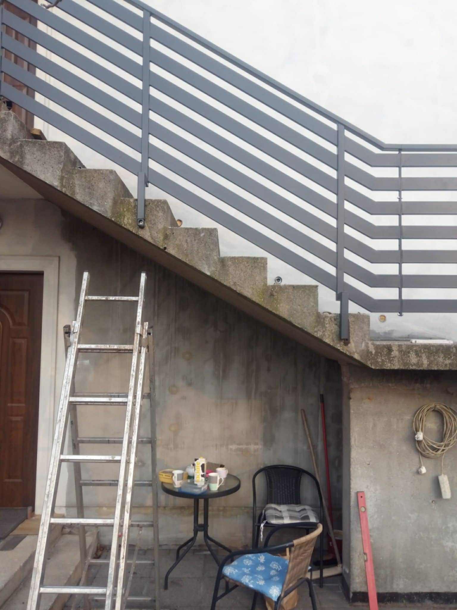 Szara, pozioma balustrada metalowa zamontowana na betonowych schodach zewnętrznych. W tle widoczna drabina, stolik z naczyniami i krzesła.