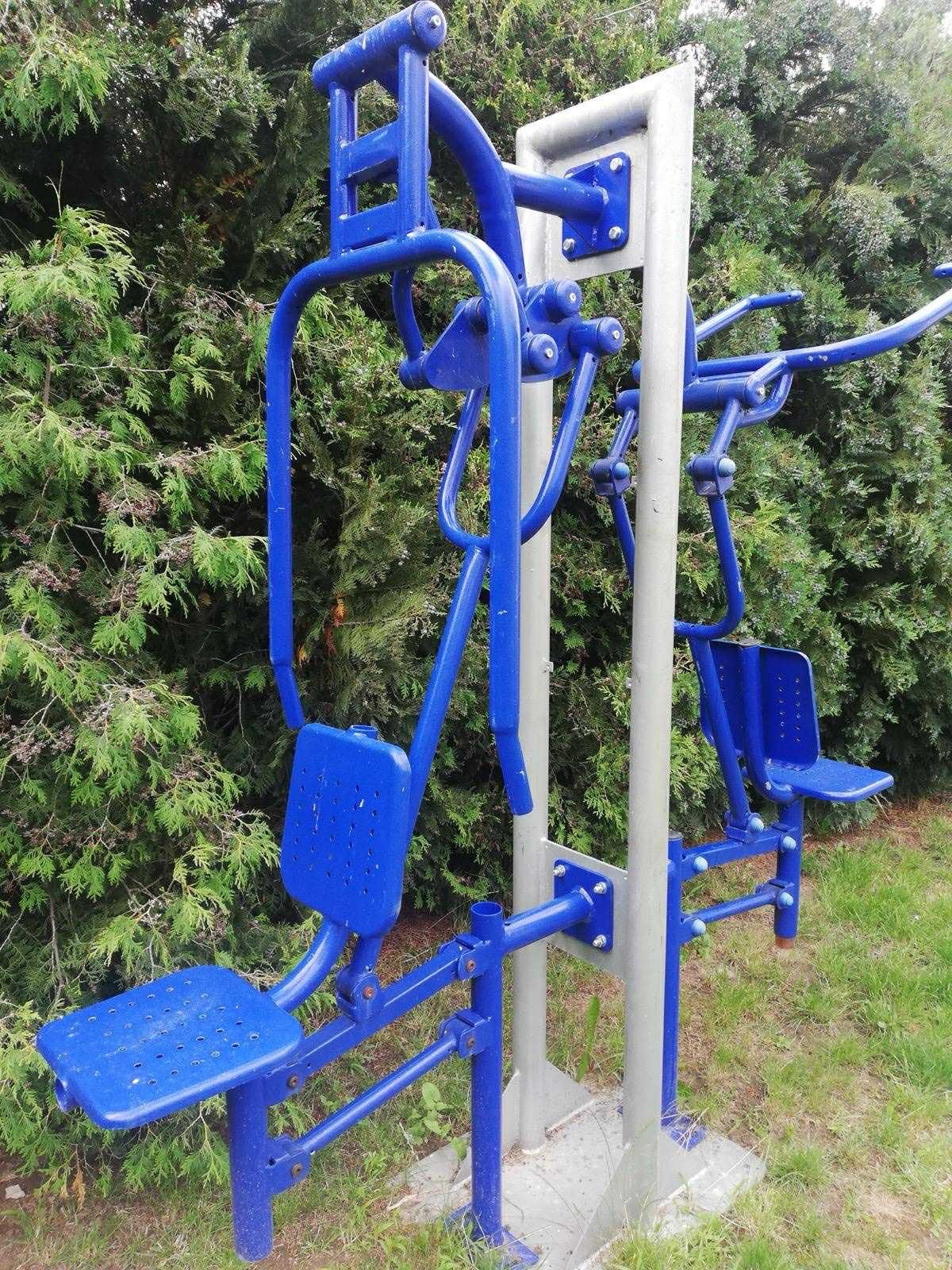 Kompleksowe, niebieskie urządzenie do ćwiczeń na świeżym powietrzu, z dwoma stanowiskami, na tle zielonych drzew i trawy. Metalowa konstrukcja z siedziskami.
