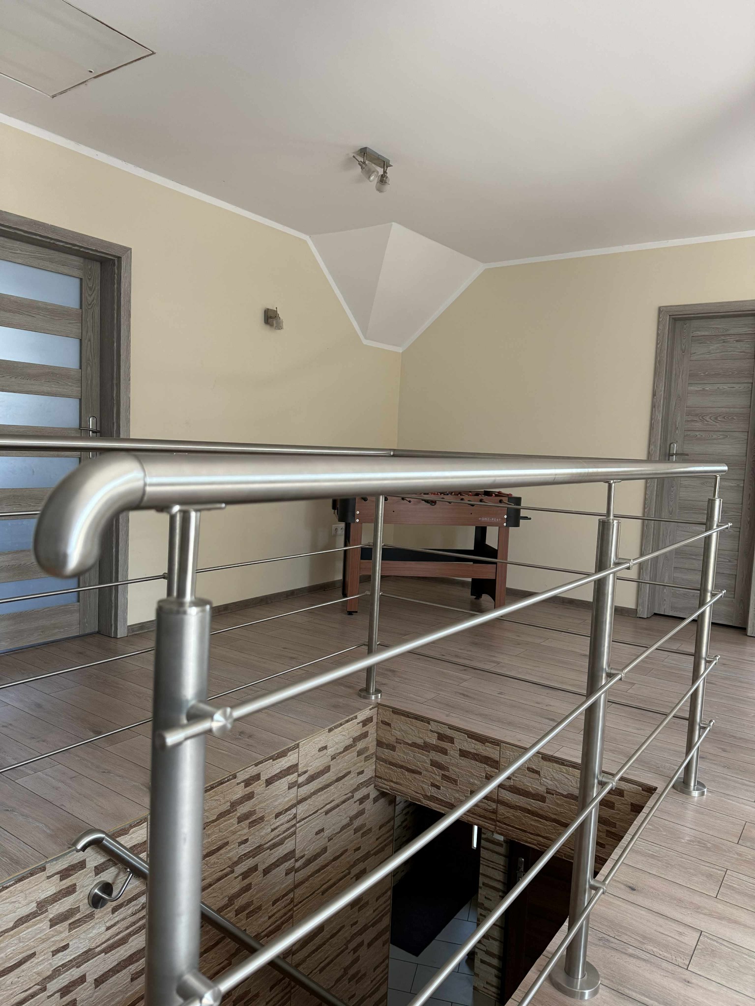 Nowoczesna balustrada nierdzewna na antresoli z widokiem na dół, z dekoracyjną okładziną ścienną. W tle widoczny stół do piłkarzyków.