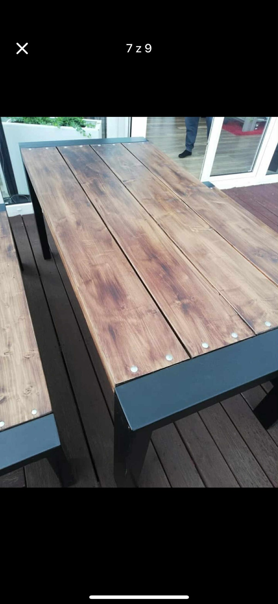 Drewniany stół ogrodowy z metalowymi nogami, widoczne śruby mocujące deski blatu. Stół na drewnianym tarasie, w tle okno.