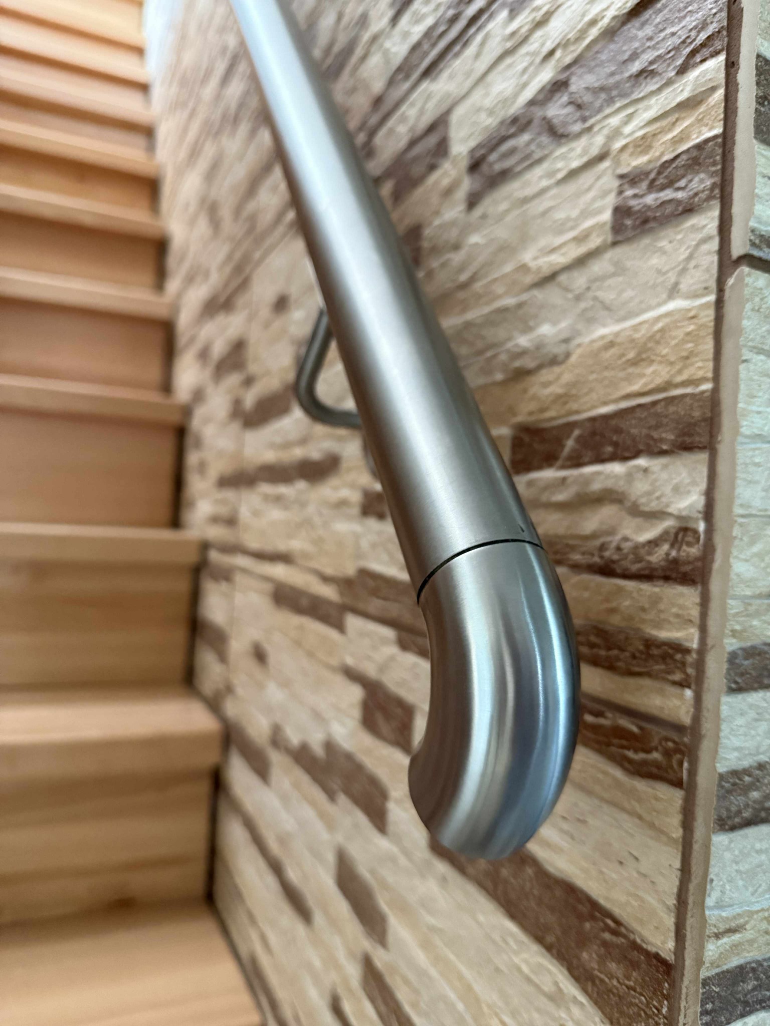 Nierdzewna poręcz schodowa, mocowana do ściany z dekoracyjną okładziną kamienną, widoczna z perspektywy dolnych stopni drewnianych schodów.
