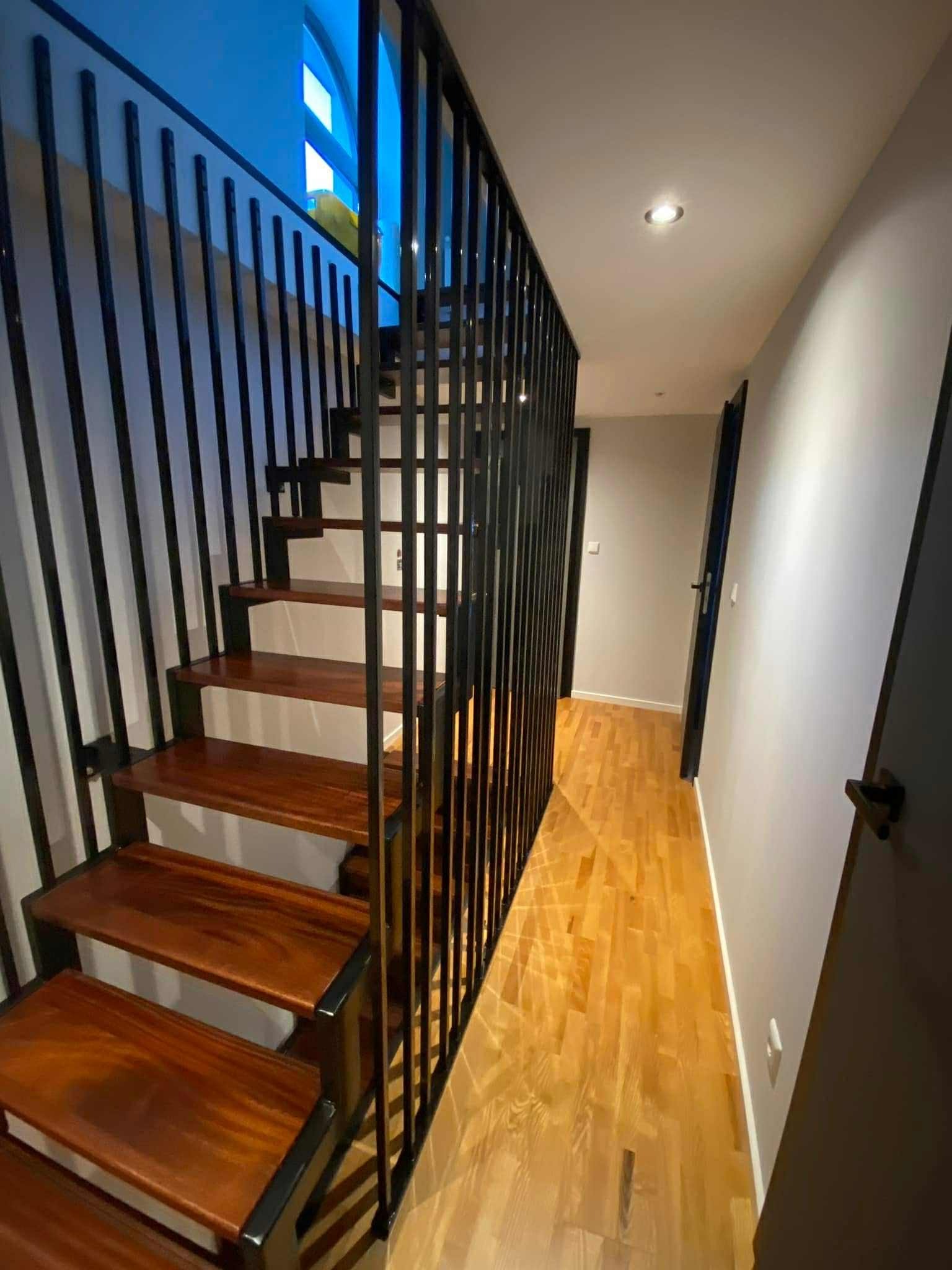 Nowoczesne schody z drewnianymi stopniami i czarną, metalową balustradą w jasnym wnętrzu. Widok na korytarz z parkietem i drzwiami.