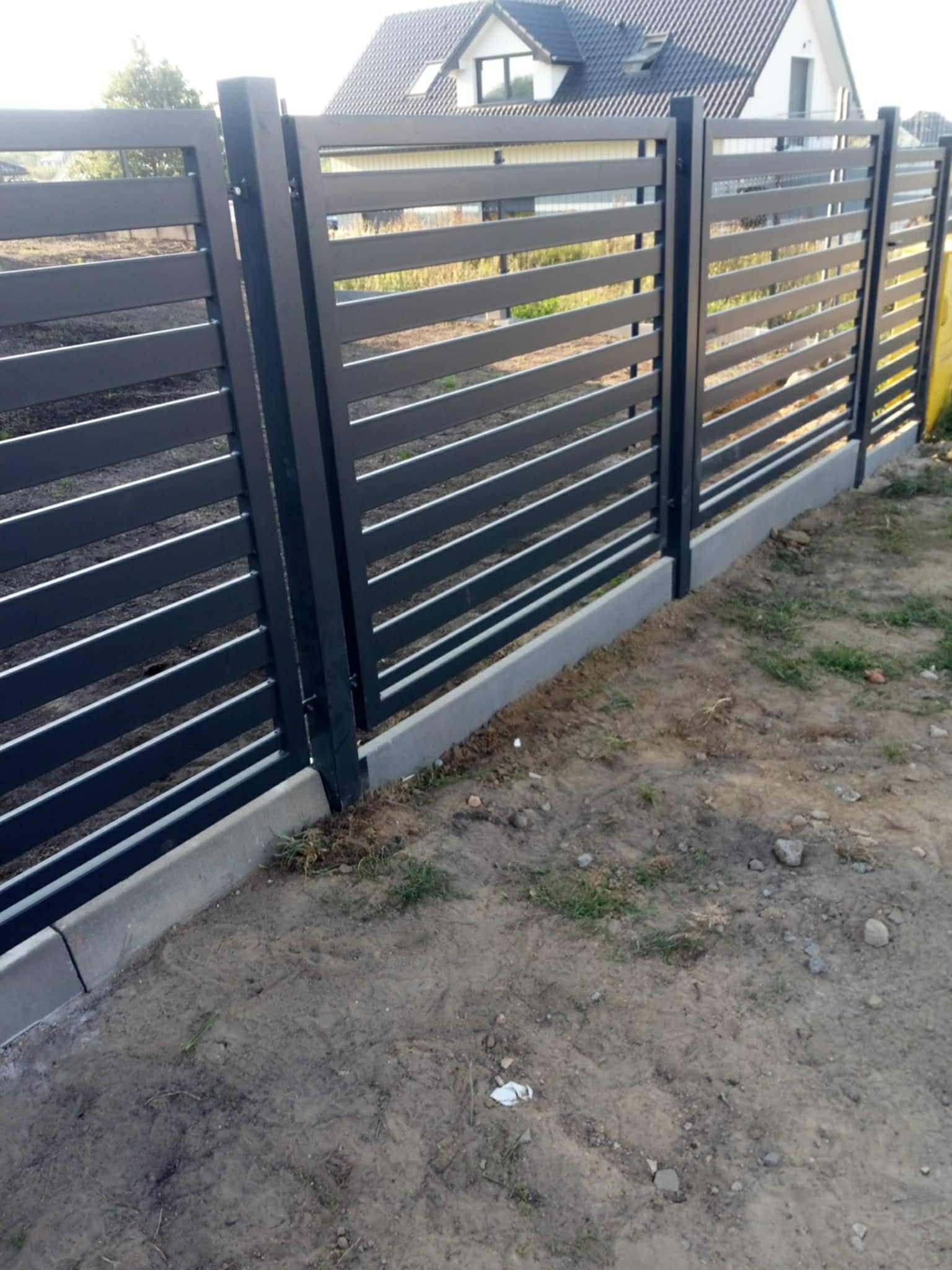 Nowoczesne ogrodzenie metalowe, poziome panele w kolorze grafitowym, osadzone na betonowej podmurówce. Dom w tle, trawa i ziemia w bliskim planie.