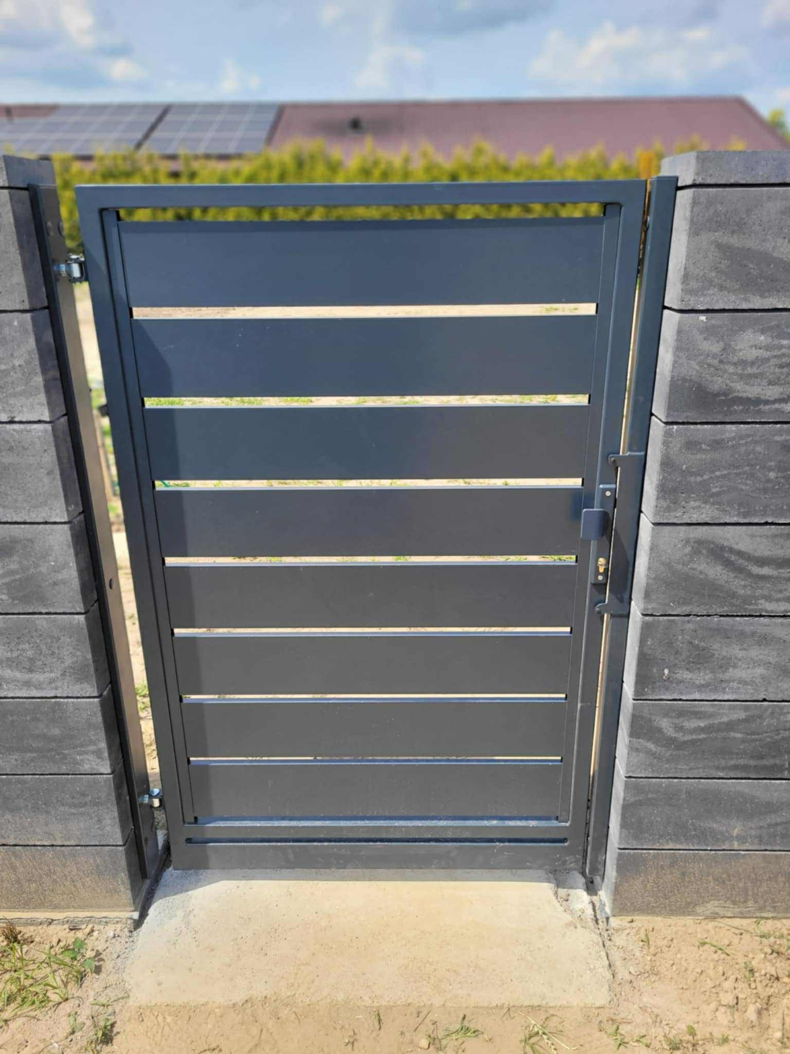 Nowoczesna, grafitowa furtka z poziomymi panelami, zamontowana między słupkami z szarej kostki. W tle widoczny dom z panelami słonecznymi na dachu.