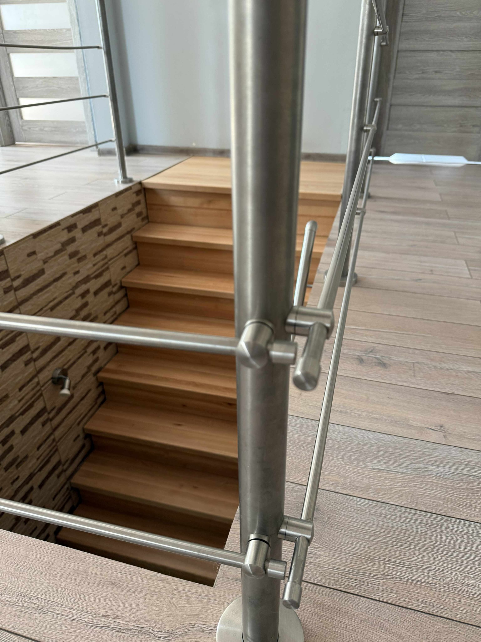 Nowoczesna balustrada nierdzewna przy drewnianych schodach, z widocznymi detalami mocowań i fakturą podłogi. Minimalistyczny design, jasne wnętrze.