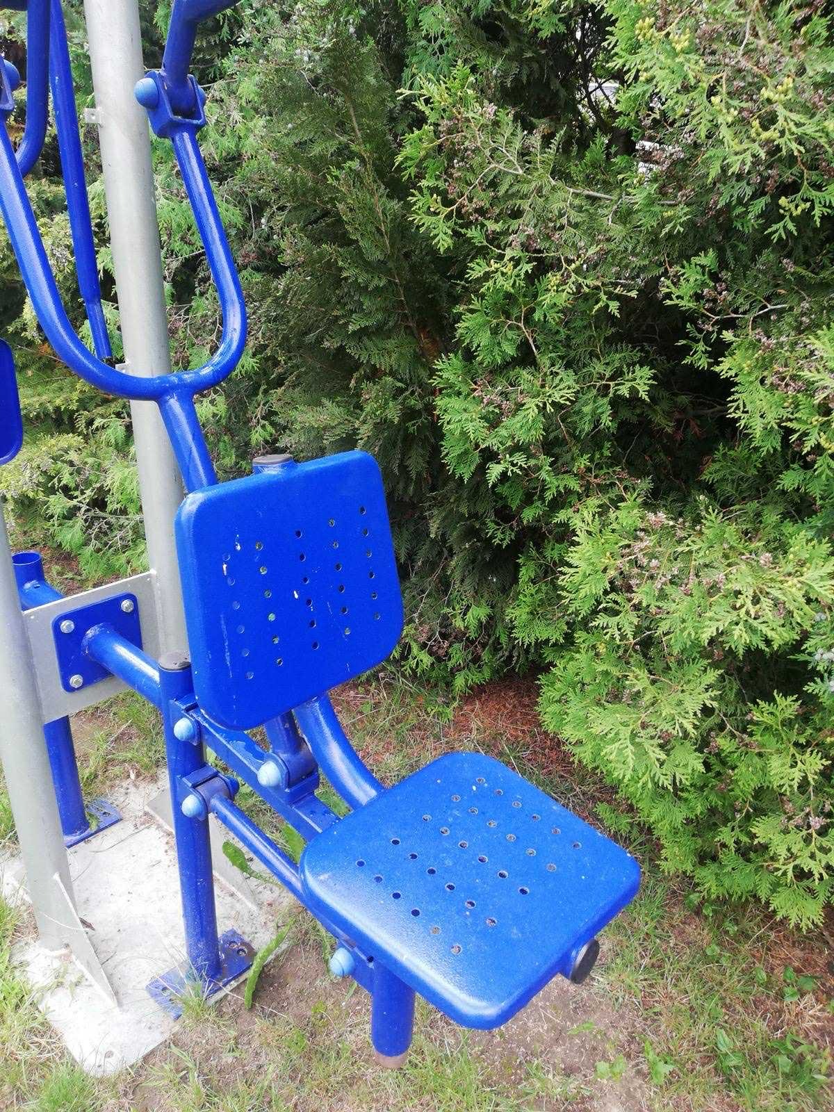 Niebieskie metalowe urządzenie do ćwiczeń na świeżym powietrzu, z perforowanym siedziskiem i oparciem, na tle zielonych krzewów. Widoczne ślady użytkowania.