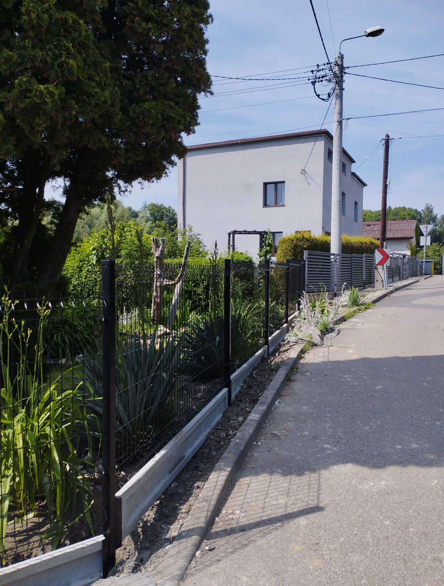 Nowoczesne ogrodzenie panelowe z podmurówką betonową, widok z ulicy, zieleń ogrodu w tle, dom jednorodzinny, słup oświetleniowy, znak drogowy.