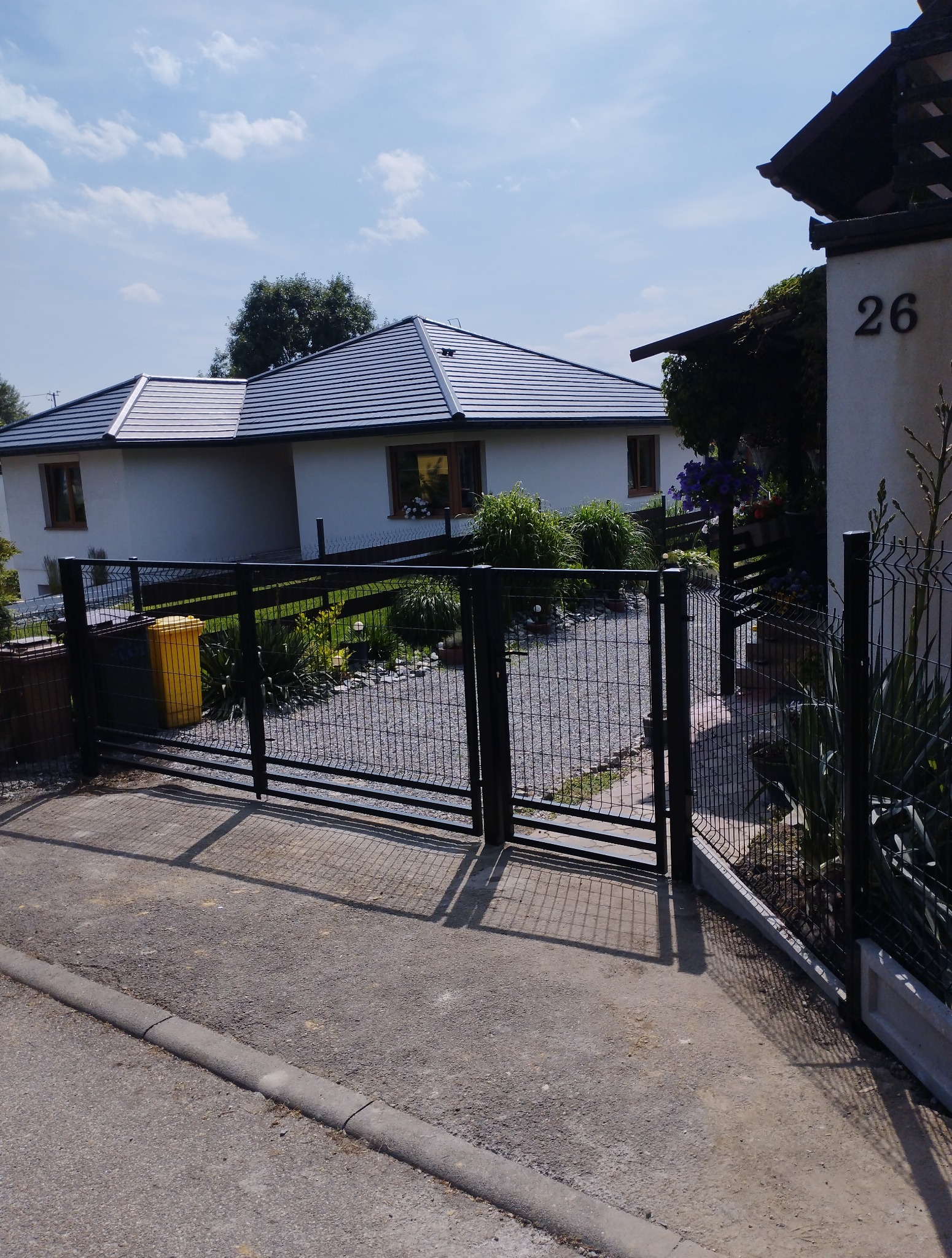 Czarna, metalowa brama z siatką na tle domu z ogrodem. Widoczny fragment chodnika i numer domu. Słoneczny dzień, brama zamknięta.