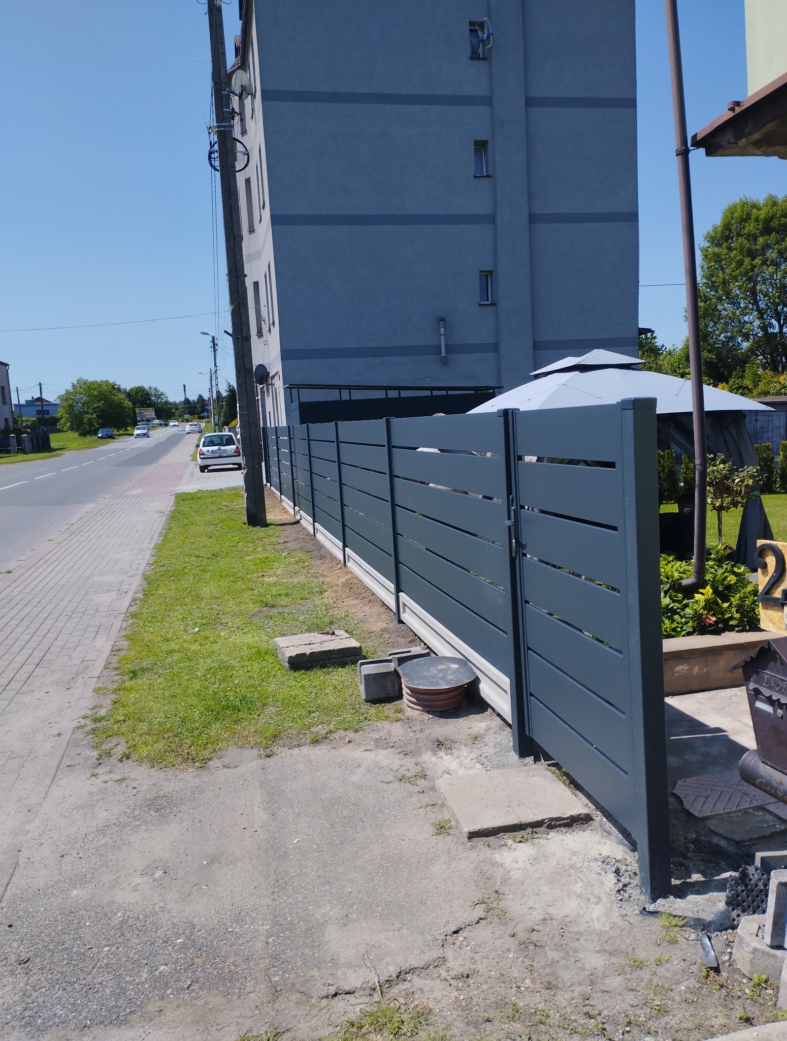 Nowoczesne, poziome przęsła w kolorze grafitowym, tworzące ogrodzenie z bramą. Widoczne betonowe podmurówki i otoczenie z chodnikiem oraz budynkiem w tle.