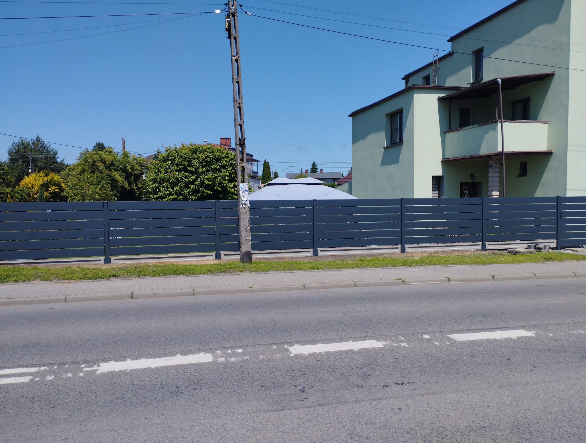 Nowoczesne, poziome, szare przęsła ogrodzeniowe na betonowej podmurówce, widoczne z ulicy na tle domu i zieleni, w słoneczny dzień.