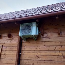 SG INSTALACJE SEBASTIAN GOSEK - Zewnętrzna jednostka klimatyzacji Fujitsu zamontowana na drewnianej ścianie domu z brązowym dachem. Widoczne mocowania i przewody instalacyjne.