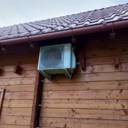 Zewnętrzna jednostka klimatyzacji Fujitsu zamontowana na drewnianej ścianie domu z brązowym dachem. Widoczne mocowania i przewody instalacyjne.