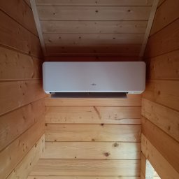 SG INSTALACJE SEBASTIAN GOSEK - Biały klimatyzator Fujitsu zamontowany w drewnianym wnętrzu, widok z dołu, ciepłe oświetlenie.