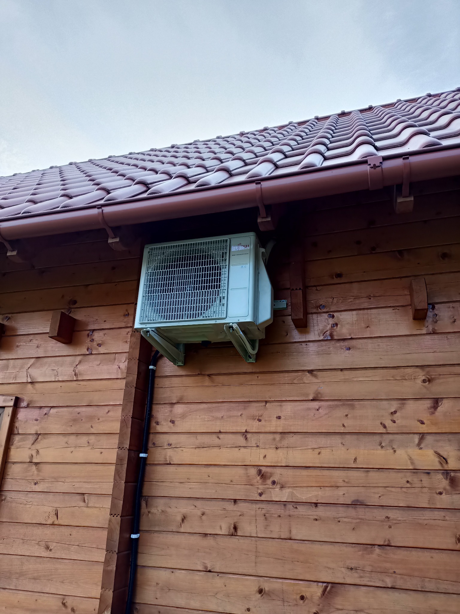 Zewnętrzna jednostka klimatyzacji Fujitsu zamontowana na drewnianej ścianie domu z brązowym dachem. Widoczne mocowania i przewody instalacyjne.