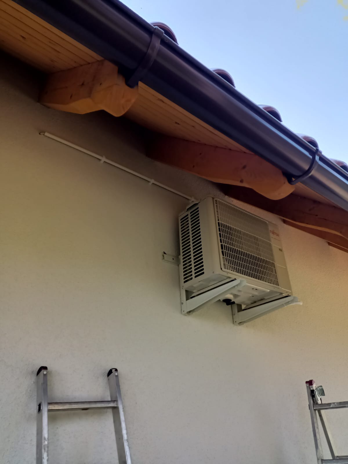 Zamontowana jednostka zewnętrzna klimatyzacji Fujitsu na elewacji budynku z widoczną rynną, drewnianym podbitką i fragmentem drabiny. Biała listwa kablowa.