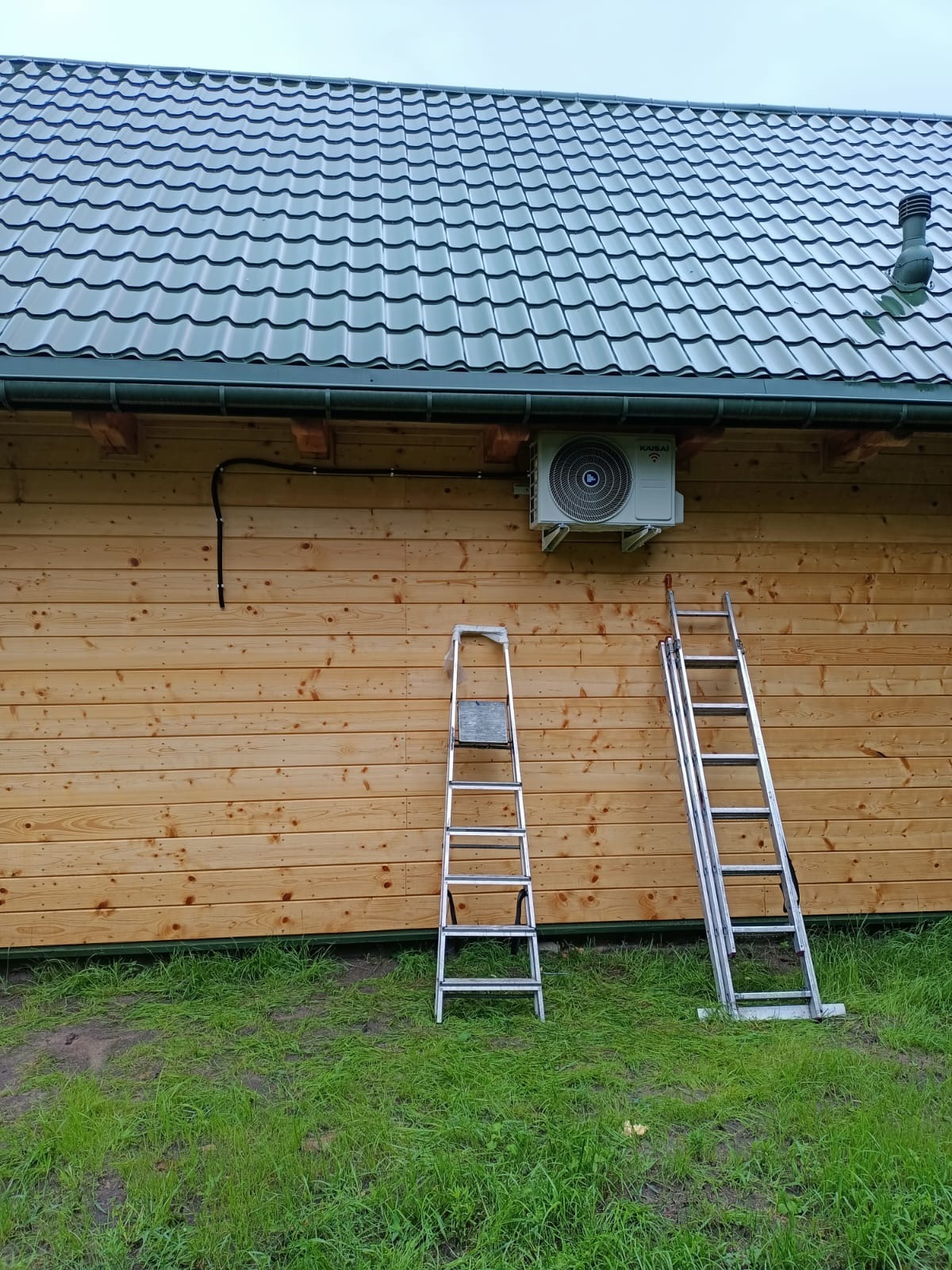 Zamontowana jednostka zewnętrzna klimatyzacji Kaisai na drewnianej elewacji domu, z dwiema przystawionymi drabinami. Widoczny fragment dachu z zieloną blachodachówką.