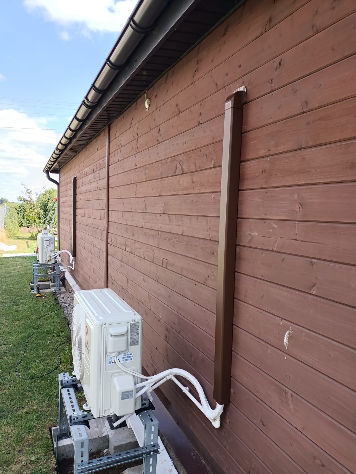Zewnętrzne jednostki klimatyzacji zamontowane na wspornikach przy drewnianej elewacji budynku. Rury odprowadzające skropliny zabezpieczone izolacją.