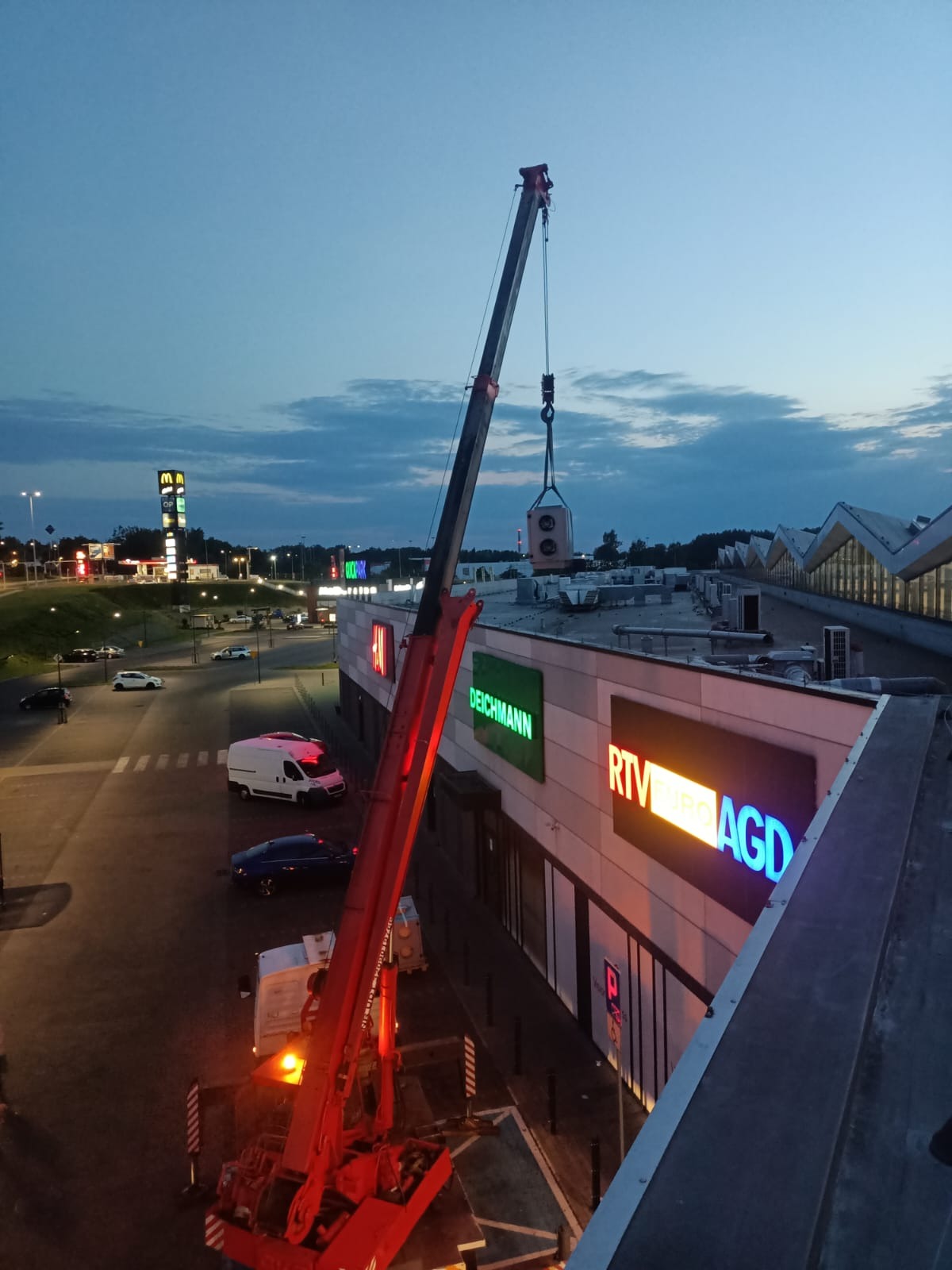 Montaż klimatyzacji na dachu centrum handlowego za pomocą dźwigu. Widoczne logo sklepów Deichmann i RTV Euro AGD. Wieczorne oświetlenie i częściowo zachmurzone niebo.