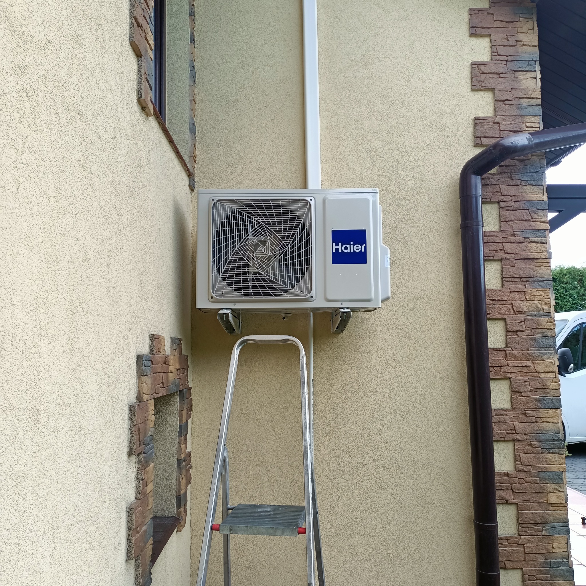 Zamontowany klimatyzator Haier na elewacji budynku z dekoracyjnym kamieniem. Widoczna drabina, biała rura instalacyjna i fragment rynny.