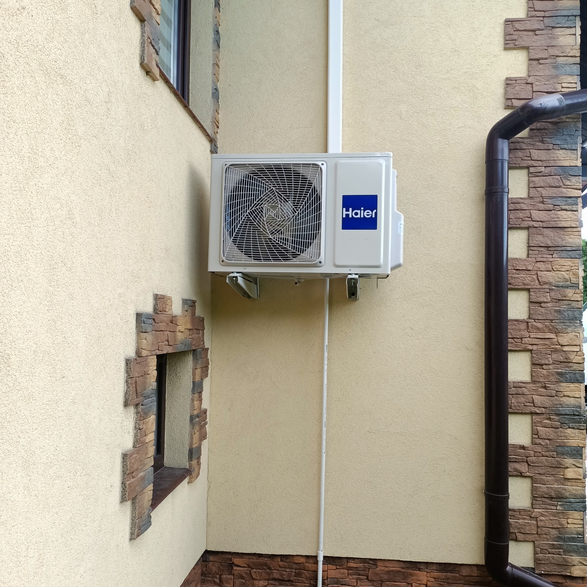 Biała klimatyzacja Haier zamontowana na beżowej elewacji budynku z dekoracyjnymi elementami z kamienia. Widoczny biały przewód instalacyjny.