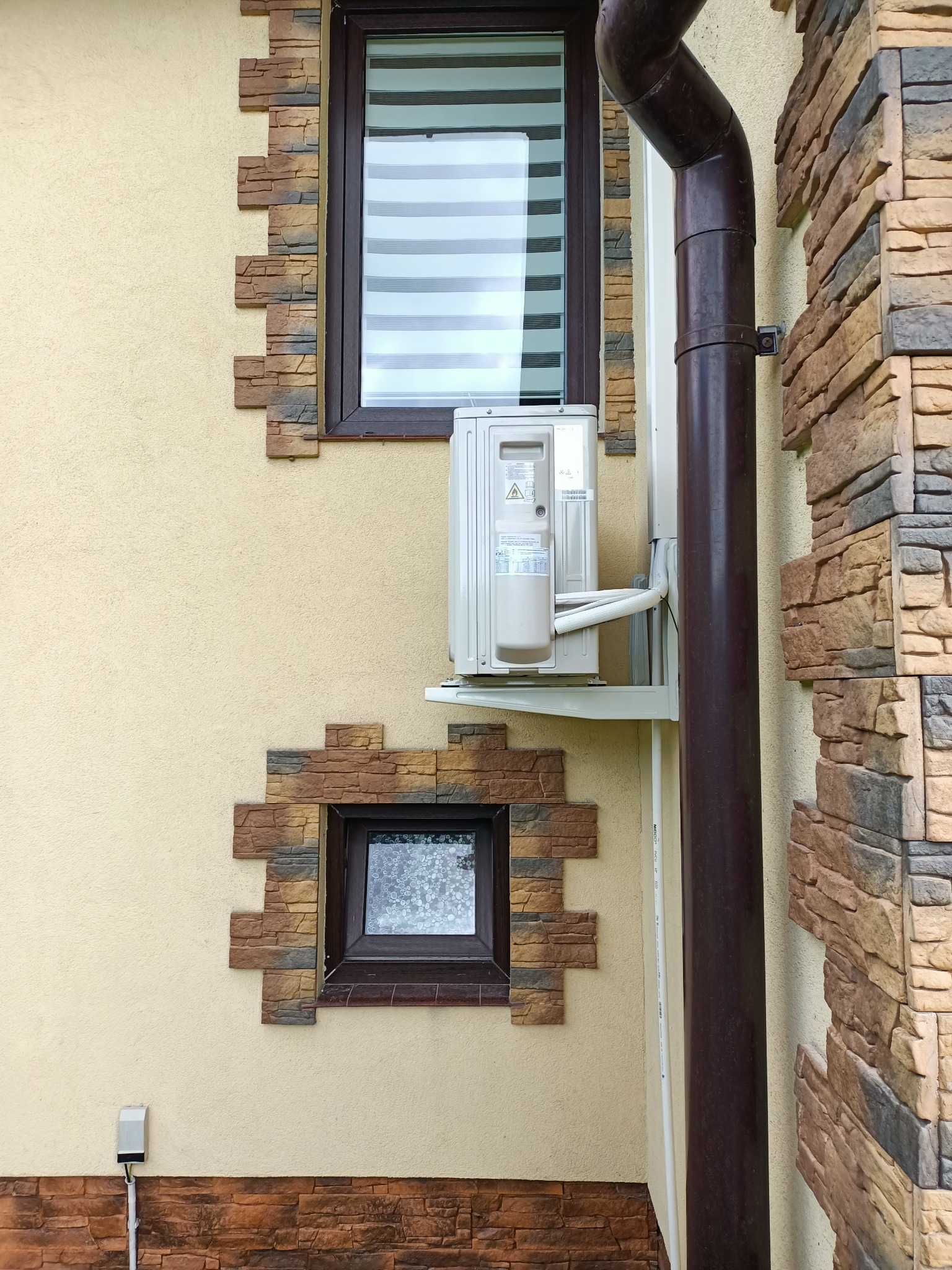 Zamontowany klimatyzator zewnętrzny na elewacji budynku z dekoracyjną okładziną kamienną i brązową rurą spustową. Widoczne okna z roletami.