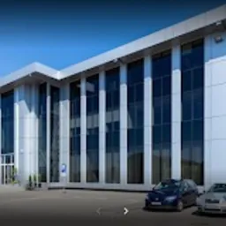 Nowoczesny, dwukondygnacyjny budynek biurowy z dużymi oknami i metalowym dachem na tle błękitnego nieba. Dwa samochody zaparkowane przed wejściem, widoczny znak parkingu.