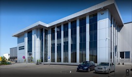 Nowoczesny, dwukondygnacyjny budynek biurowy z dużymi oknami i metalowym dachem na tle błękitnego nieba. Dwa samochody zaparkowane przed wejściem, widoczny znak parkingu.