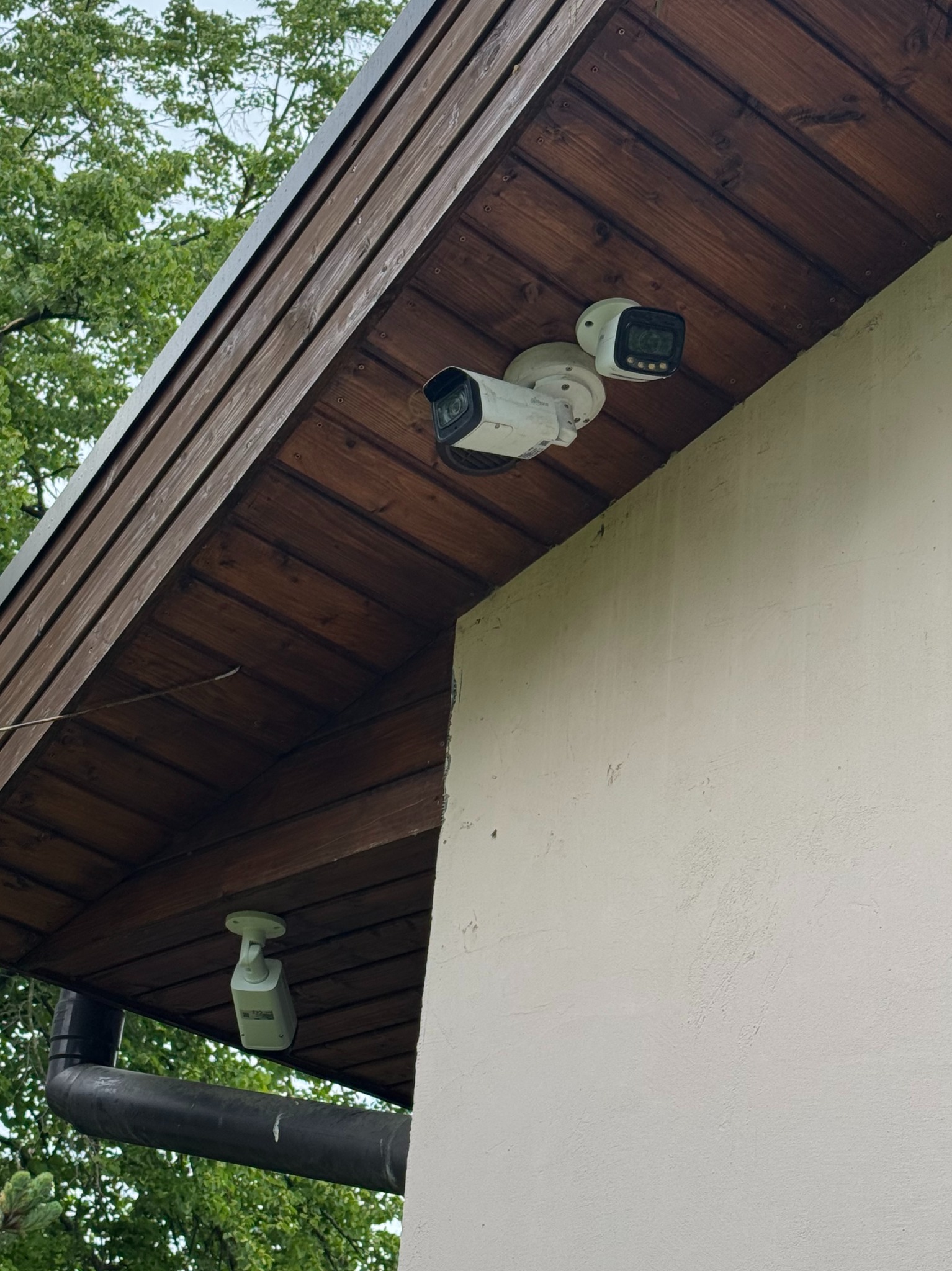 Zewnętrzne kamery monitoringu przymocowane do drewnianego podbitki dachu budynku z jasną elewacją. Widoczne drzewa w tle. Ujęcie z dołu.