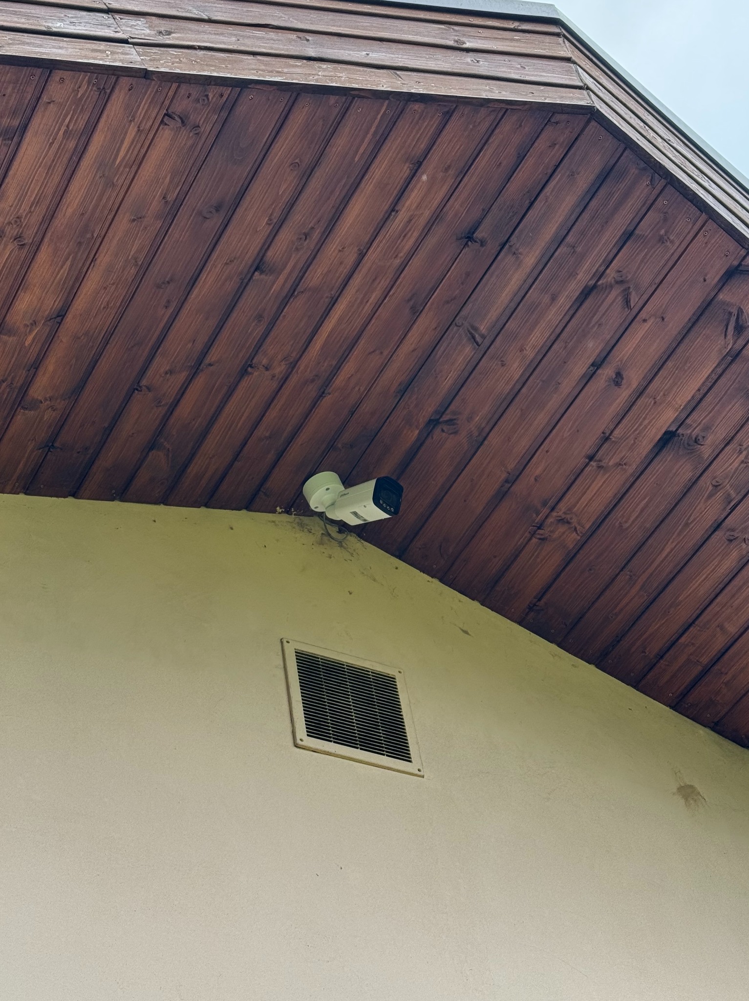 Kamera monitoringu IP zamontowana pod drewnianym okapem w Koninko, widoczna kratka wentylacyjna na elewacji budynku. Ujęcie z dołu, zbliżenie.