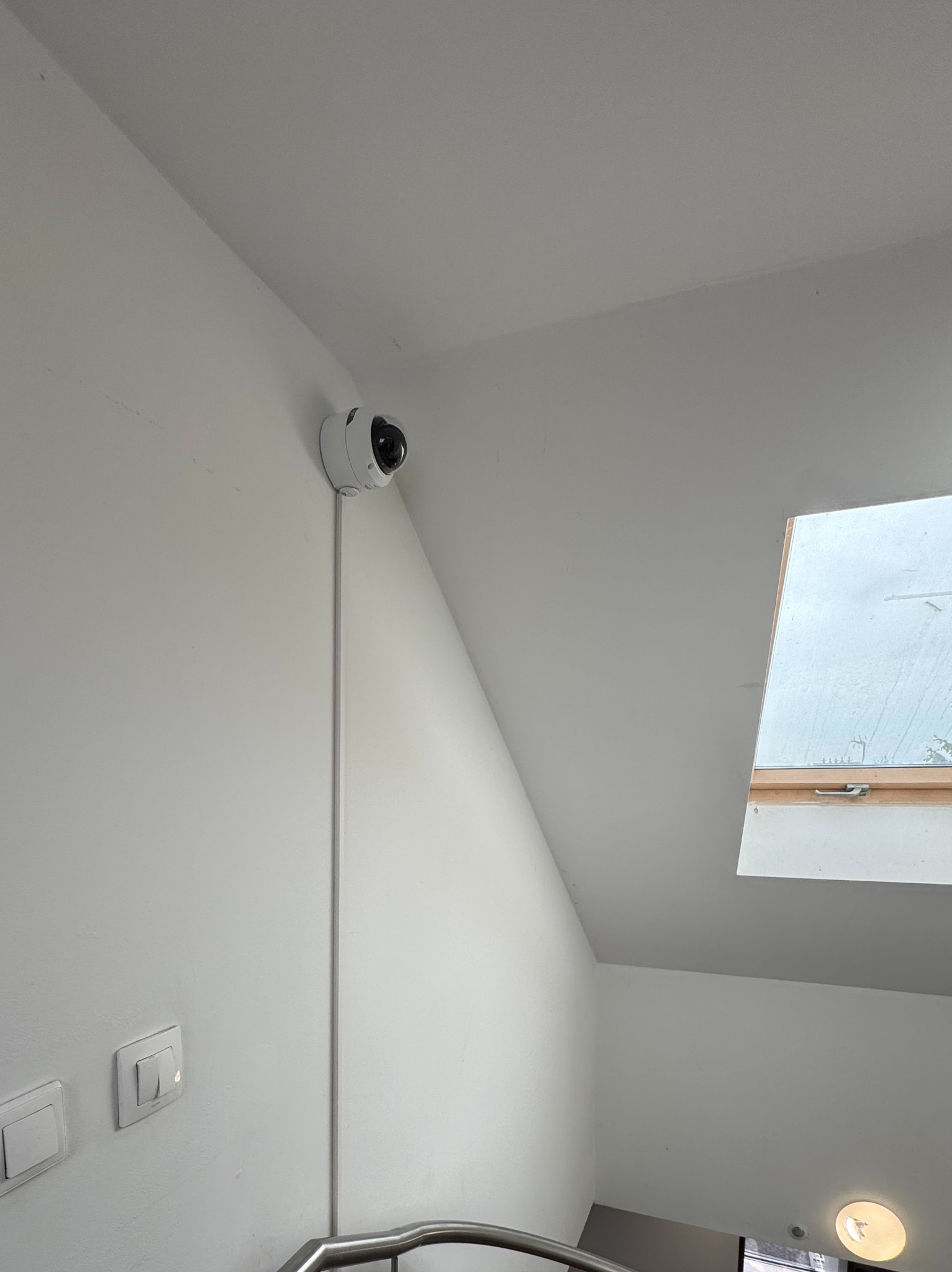 Kamera monitoringu zamontowana na białej ścianie wewnątrz budynku, widoczne okno dachowe i fragment schodów. Kabel estetycznie poprowadzony wzdłuż ściany.
