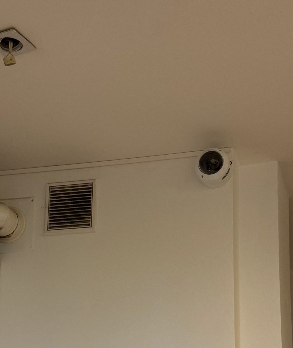 Kamera kopułkowa, biała, zamontowana w rogu pomieszczenia, widoczna kratka wentylacyjna i element instalacji pod sufitem. Oświetlenie dzienne.