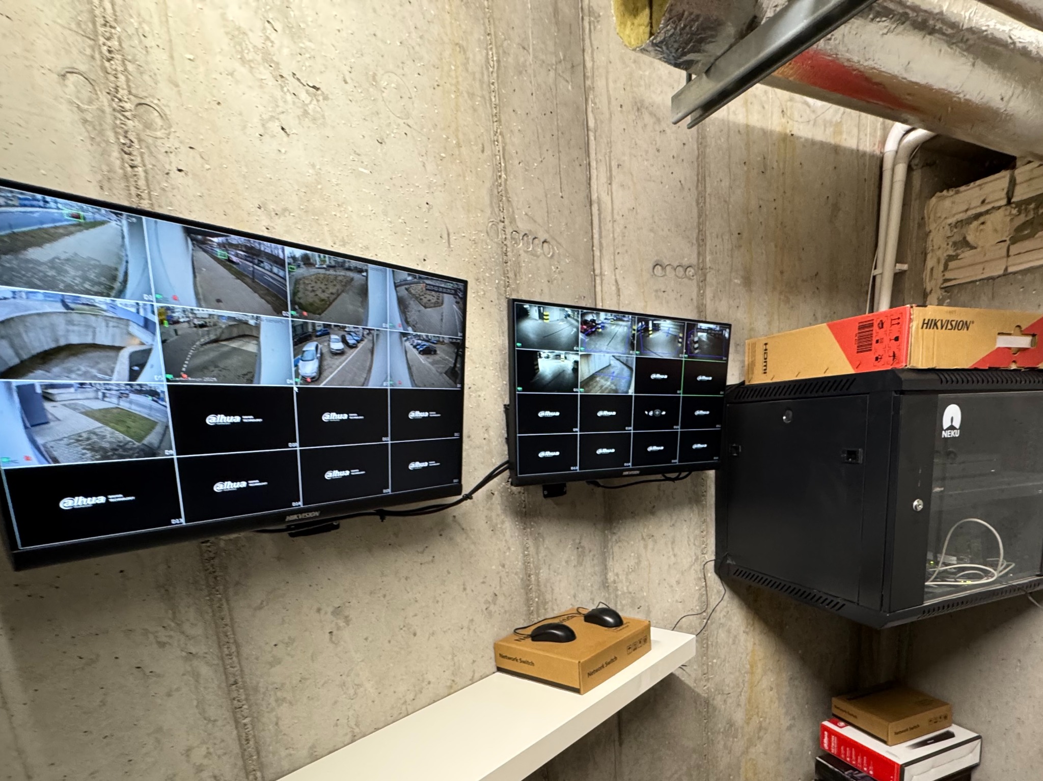 Dwa monitory Dahua prezentują obraz z kamer monitoringu. Obok szafa rack NEKU z urządzeniami Hikvision. Widok na surową betonową ścianę.