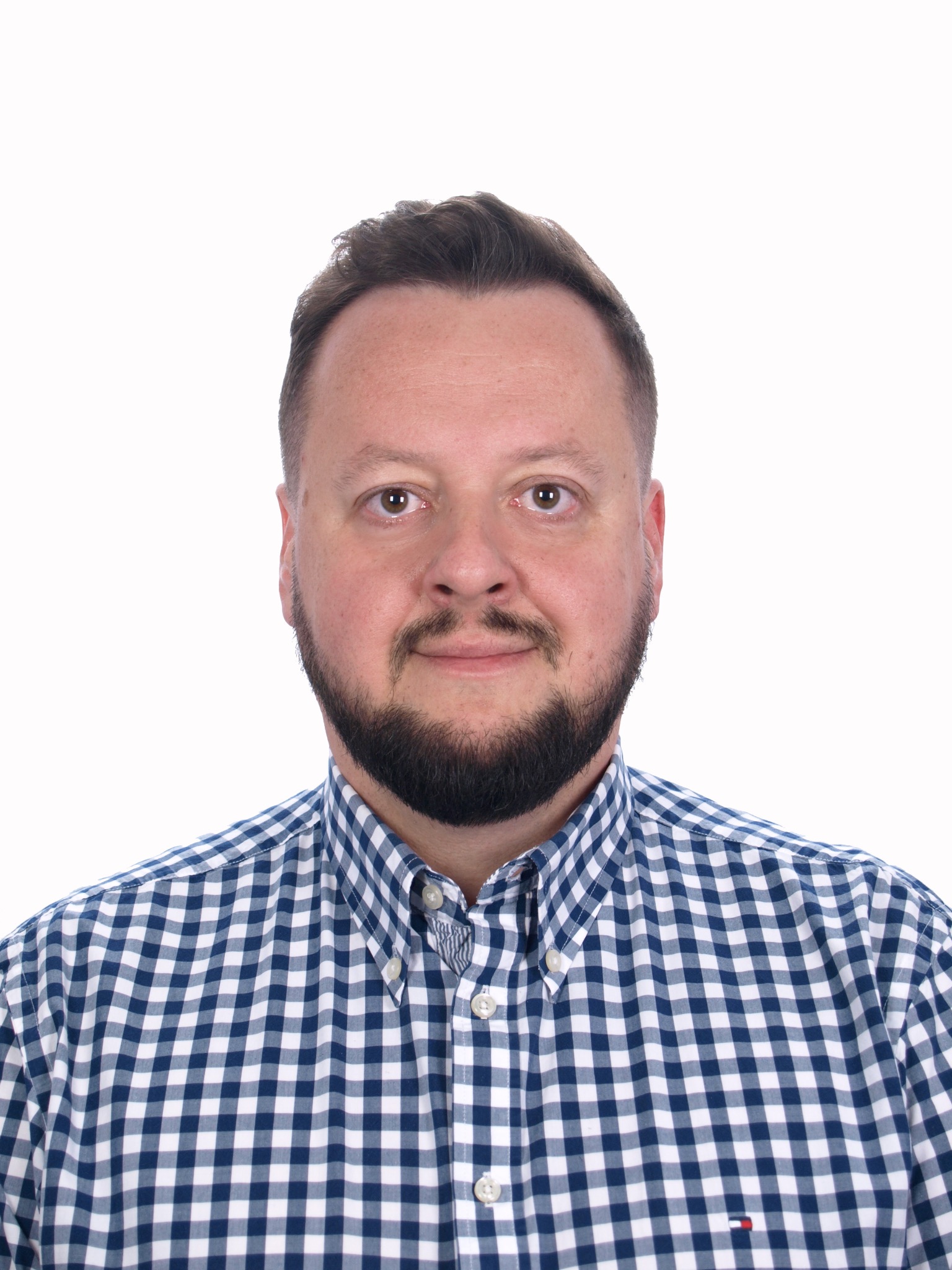 Portret mężczyzny z brodą w niebiesko-białej koszuli w kratę na jasnym tle, uśmiechający się i patrzący wprost w obiektyw.