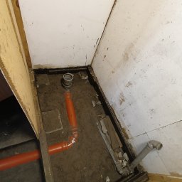 Fuszkin - Fragment podłogi z instalacją kanalizacyjną w trakcie montażu. Widoczne rury PCV w kolorze pomarańczowym i szarym. Podłoże ziemne, ściany w stanie surowym.