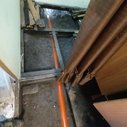 Fuszkin - Pomarańczowa rura kanalizacyjna ułożona w wykopie pod podłogą, widoczne deski i fragment zasłony. Prace instalacyjne w trakcie remontu.