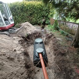 Fuszkin - Montaż szamba betonowego w wykopie, widoczny fragment rury odprowadzającej i koparka w tle. Teren zielony z żywopłotem.