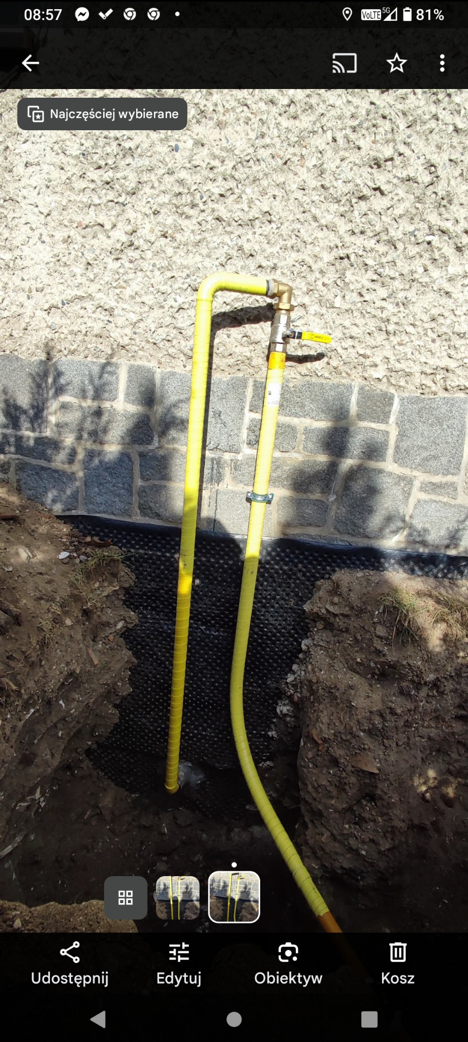 Żółta rura gazowa z zaworem, częściowo zakopana w ziemi przy ścianie budynku. Widoczne zabezpieczenie ściany folią kubełkową. Kąt ujęcia z dołu.