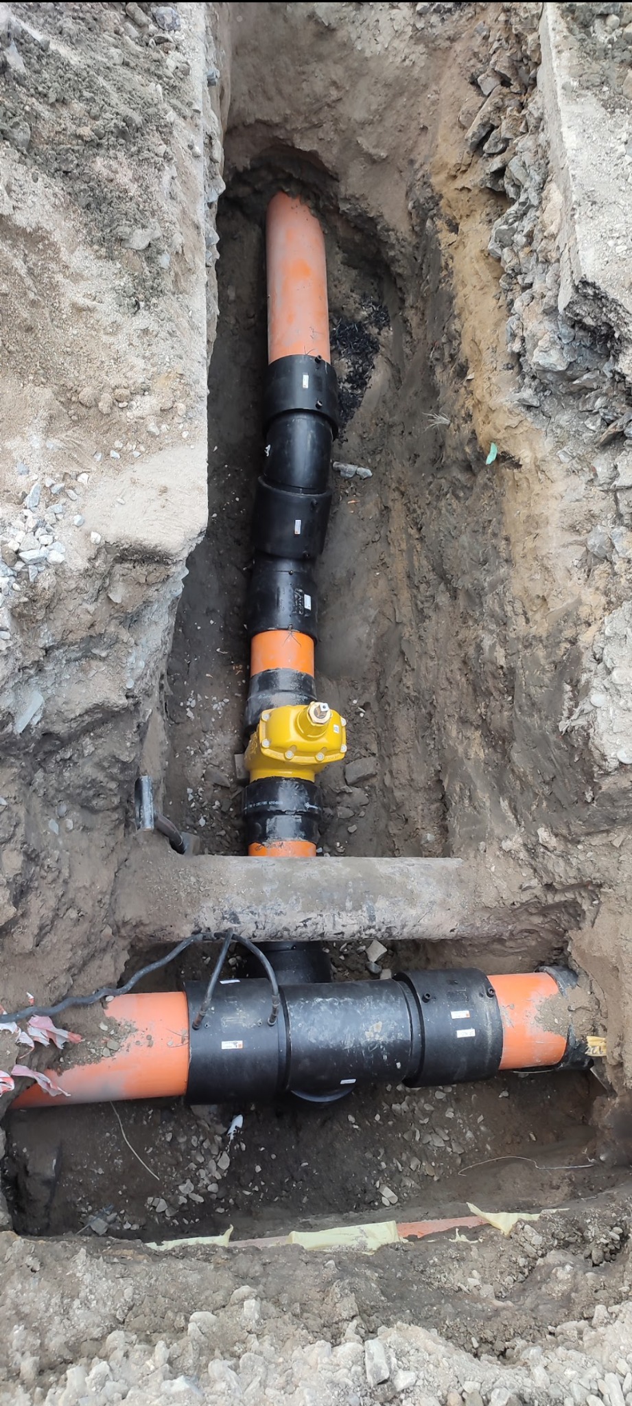 Pomarańczowe i czarne rury kanalizacyjne w wykopie ziemnym, z żółtym zaworem. Widoczne złącza i fragmenty przewodów elektrycznych.