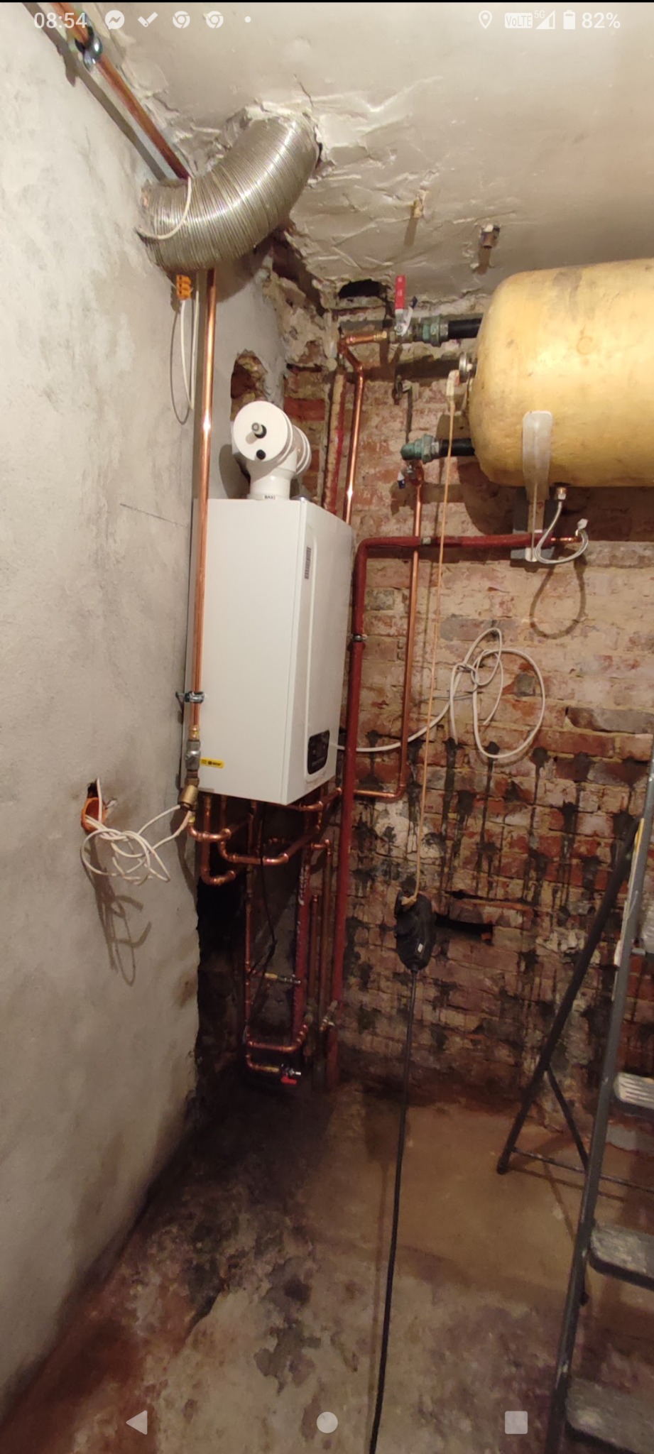 Biały piec gazowy BAXI z miedzianymi rurami w surowym pomieszczeniu z odsłoniętą cegłą i drabinką. Widoczny zbiornik na wodę i przewód wentylacyjny.